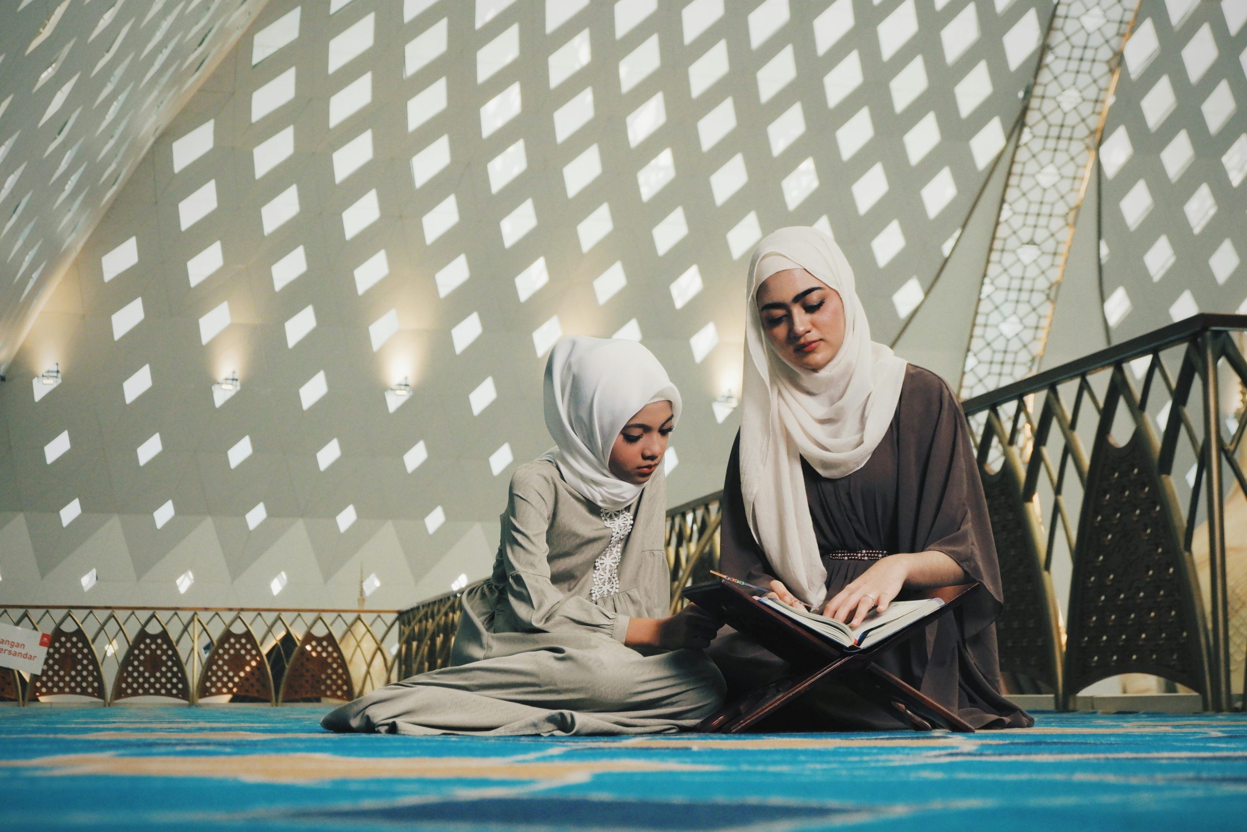 A Muslim woman and her daughter reading the Quran together inside a beautifully lit mosque.