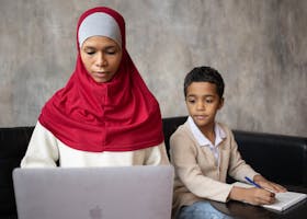 Muslim pensive ethnic mother typing on netbook while son with pen taking notes in notebook